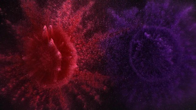 In real slow motion footage against a black background, a red pile and a purple pile of powder with a sunburst of powder explode like fireworks and begin to fall. Top shot.