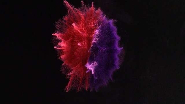 In real slow motion footage against a black background, a two color circular pile of powder bursts with the red on the left and purple on the right while still maintaining color separation. Top Shot.