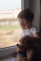 Stay at home quarantine coronavirus prevention of the pandemic. A child and his Teddy bear on the windowsill and look out the window. The view of the street. Prevention of the epidemic.