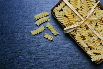 Background and space for text with a cardboard box of raw spiral noodles photographed from above, on a gray slate board and a bow made of bast