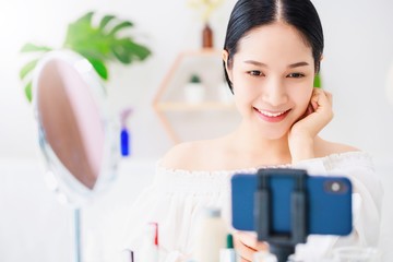 Beautiful asian woman blogger is showing how to make up and use cosmetics. In front of the mirror and smartphone recording vlog video live streaming at home.Skincare for healthy face concept.