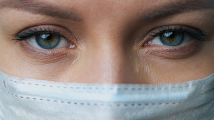 A female doctor therapist in a white robe, mask and gloves. Face close-up. The doctor cries and prays. Tears in eyes. Pandemic and virus epidemic. Coronavirus covid-19.