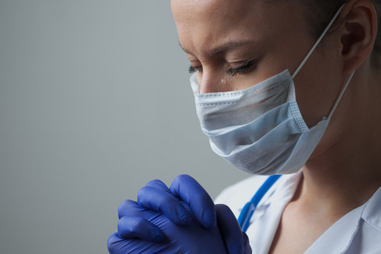A female doctor therapist in a white robe, mask and gloves. Face close-up. The doctor cries and prays. Tears in eyes. Pandemic and virus epidemic. Coronavirus covid-19.
