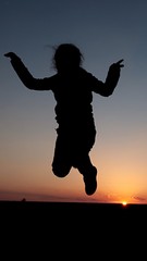 Fototapeta na wymiar Silhouette of small girl jumping with raised hands in sunset above spring field. 