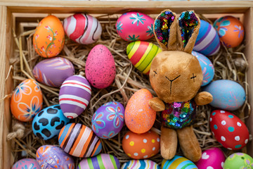 Fototapeta na wymiar flat lay of colorful vibrant easter eggs with stripe flower pattern paint, laying on basket nest with brown bunny rabbit soft toy, blue wooden table background representing celebrating easter holidays