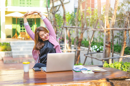 Asian Businesswoman Stretching With Both Of Her Arms Stretched Up In The Air With Back Straight, And Getting Tired From Working On The Laptop Computer, Calculator, Notebook And Clipboard On The Table