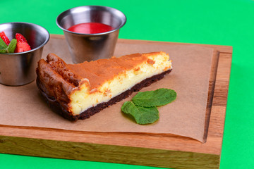 A piece of sweet cheesecake with fresh raw strawberry and sauce served on rustic wooden board over green background.