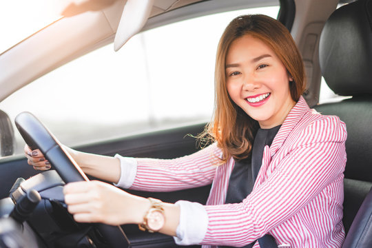 asian businesswoman driving a car holding a steering wheel with both hands while smiling joyfully, sitting in the driver seat with seatbelt on wearing suit and with sunset light shining in background