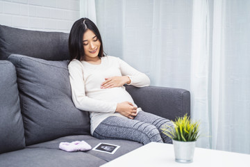 beautiful asian pregnant woman placing hands on baby lump feeling heartbeat smiling joyfully, sitting on sofa relaxing resting from tiredness, in living room with brick texture wall and white curtains