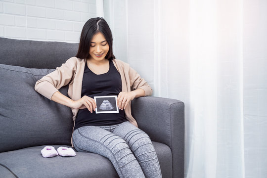 beautiful asian pregnant woman sitting on sofa holding ultrasound baby picture while placing hand on belly resting relaxing in living room from hormone stress, wearing comfy stretch pants and cardigan