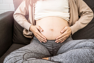 Close up asian pregnant woman placing hands on baby lump stomach feeling baby heartbeat, sitting on sofa relaxing and resting from tiredness, in living room with brick texture wall and white curtains