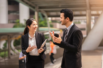 asian businesswoman holding a laptop talking and laughing joyfully with a caucasian businessman holding a phone, talking about business work or socializing, within urban city structure in background..