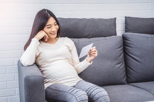 beautiful asian pregnant woman sitting on sofa holding ultrasound baby picture while placing hand on belly, resting relaxing in living room from hormone stress, wearing comfy stretch pants and jumper