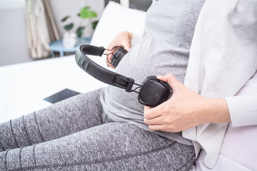 Close up asian pregnant woman holding and using black headphone to her pregnant belly, letting the baby listening inside, smiling with happiness siting leaning on white wooden bed relaxing in bedroom