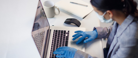 Young woman employee wearing protective gloves on hands and mask on face working from office or home using laptop on desk. Preventing corona virus covid-19 spread during epidemic outbreak