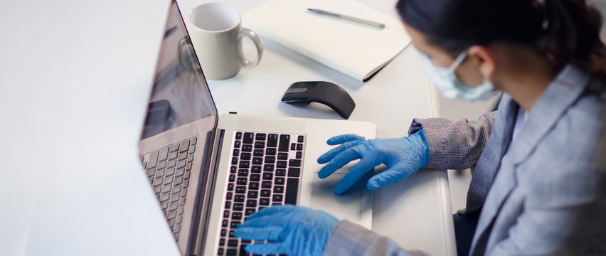 Young woman employee wearing protective gloves on hands and mask on face working from office or home using laptop on desk. Preventing corona virus covid-19 spread during epidemic outbreak