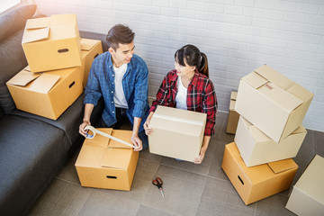 asian couple moving house packing belongings, objects and item away in a brown cardboard box, in the living room helping each other packaging and stacking boxes, smiling joyfully and working together