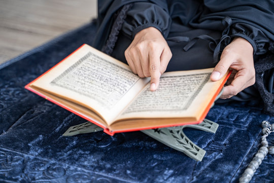 close up asian muslim woman reading the quran resting on rahel stand and using finger pointing along, sitting on vintage prayer mat on knees, with misbaha resting on matt, bright cool tone background