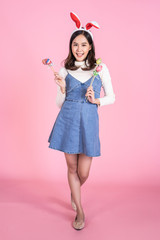 beautiful cute asian girl wearing bunny head band and denim overalls smiling playfully at camera and holding bouquet of easter eggs against chest celebrating easter holiday. isolated pink background