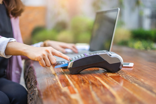 Asian Businesswoman Using And Pressing A Green Button On A Payment Machine With A Credit Card Inserted Within It, With Smartphone, Calculator Pen And Laptop Computer Laid Together On A Wooden Table