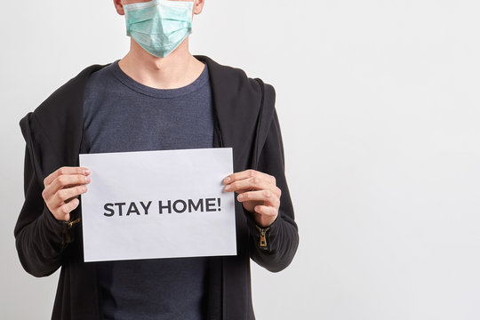 A man wearing protective medical face mask during coronavirus outbreak and keeps in hands a paper sheet with inscription STAY HOME. Virus spread flu prevention carantine. Light background