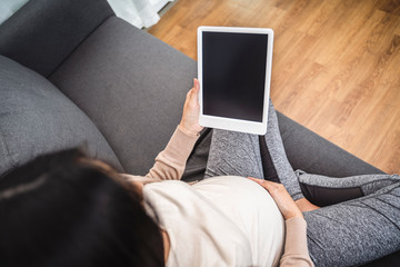 beautiful asian pregnant woman smiling using smart tablet device watching video placing hands on baby lump, sitting on sofa relaxing resting in living room with brick texture wall and white curtains