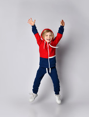 Active happy laughing blond kid boy in blue and red sportswear hoodie and pants jumps with his hands up