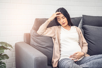 beautiful asian pregnant woman feeling stress from high pregnancy hormones, headache and morning sickness placing hand on forehead with closed eyes, sitting on sofa relaxing resting in living room