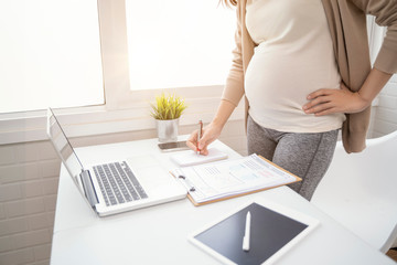 pregnant asian woman working from home in modern white office using pen writing on notepad with tablet laptop computer technology smiling with happiness of motherhood pregnancy expectant new born baby