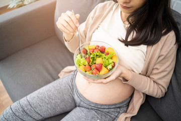 Beautiful asian pregnant woman holding a bowl of salad of vegetables and fruits, using a fork eating tomato. Eating healthy having lunch sitting on the grey sofa, relaxing and resting living room