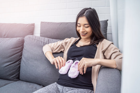 beautiful asian pregnant woman sitting on sofa holding pink fury fluffy baby shoes placing hand on belly, resting relaxing in living room from hormone stress, wearing comfy stretch pants and cardigan