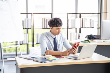 handsome asian male worker using mobile phone contacting colleagues, siting at desk with office equipment and devices such as laptop computer, books and pens, representing contact us in work area