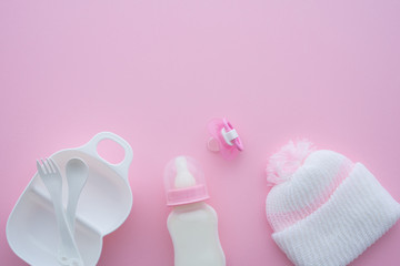 Top view of baby toddler feeding bottle with milk, knit winter hat, pacifier or soother and grinder bowl on pink background. Newborn accessories prepare for baby girl or toddler, breastfeeding concept
