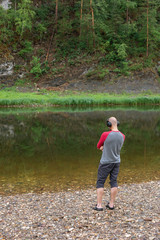 Fototapeta na wymiar Back view of young hipster man listening to music in headphones on river bank. Far from big city bustle. Relaxing outdoors. Melomaniac