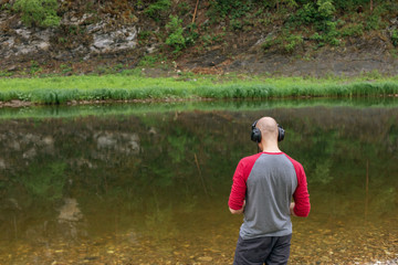 Fototapeta premium Rear view of bald young man standing on river bank and listening to music in headphones. Far from big city bustle. Relaxing outdoors. Melomaniac