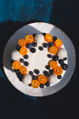 A top of a homemade cake covered with white cream and decorated with fresh blueberries, cut kumquats and coconut sweets. Served on a white handmade plate on blue background. 