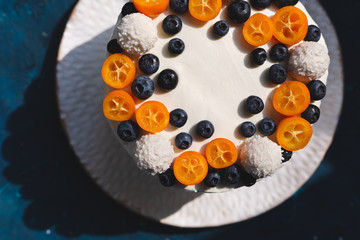 A top of a homemade pie covered with white cream and decorated with fresh fruits, berries and coconut sweets. Served on a white plate on blue background. Cut view. Natural sun lighting.