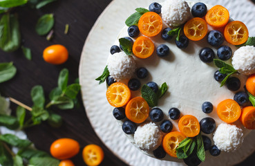 Top view of a delicious homemade birthday or wedding cake with white curd cream and decorated with fresh berries, kumquat and mint leaves on a big white plate on dark wooden background. Cut view