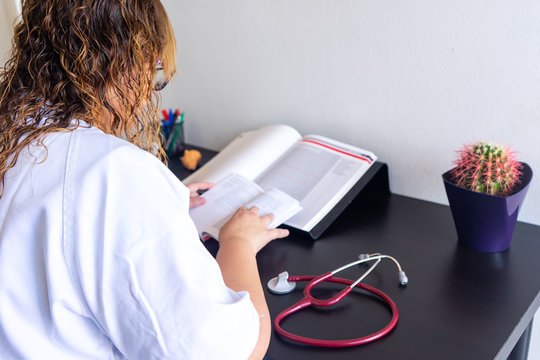 Young woman studying medicine sitting at a desk. There is also a cardiology textbook where you can see electrocardiograms.  On the desk there is a stethoscope and a cactus. 