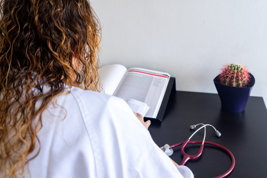Young woman studying medicine sitting at a desk. There is also a cardiology textbook where you can see electrocardiograms.  On the desk there is a stethoscope and a cactus. 