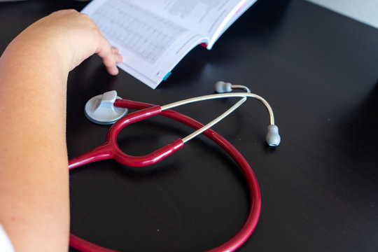 On a desk in the foreground a stethoscope. In the background is a pencil with pens, a cactus and a laptop. All out of focus.