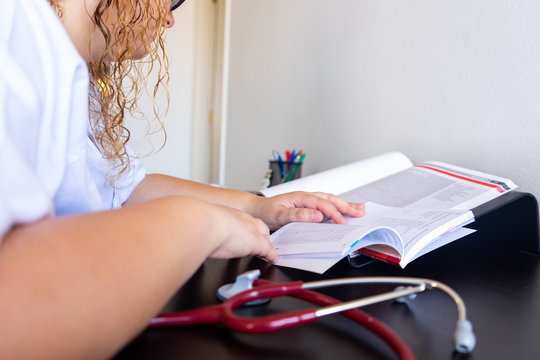 Young woman studying medicine sitting at a desk. There is also a cardiology textbook where you can see electrocardiograms.  On the desk there is a stethoscope and a cactus. 