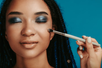 Calm peaceful attractive afro american woman with closed eyes on photo. Hand hold make up brush and touch model's cheek. Eyeshadows with blue metal color. Isolated over blue background.