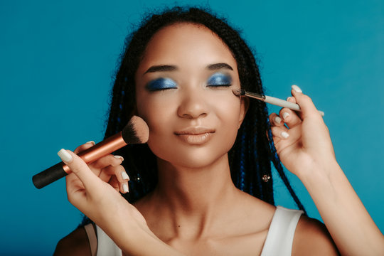 Portrait of young afro american woman isolated over blue background. Caucasian hands with two make up brushes close to model's face. Eyeshadows with blue metal color.