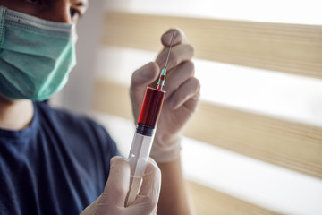 Young man with mask and gloves looking at injection full with blood. Be responsible, stay at home. World pandemic.