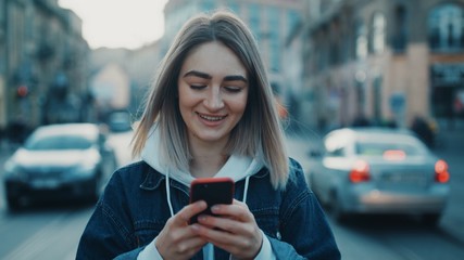 Slim pretty girl walks trought the city holding smartphone writing texting a message on social media looking around road close up