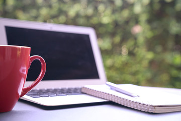 Computer laptop with notepad and coffee in red cup with green nature background. Work remotely or from home.