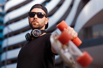 Young man with headphones around neck wearing hat and sunglasses holding a skateboard in his hand and posing in the city. © djile