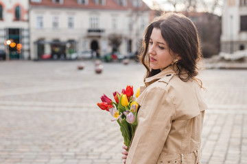 Fototapeta premium Beautiful young woman with spring tulips flowers bouquet at city street. Happy girl walking outdoors. Spring portrait of pretty female in old town