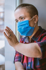 A man in a medical mask sits by the window of a house in isolation during a coronavirus pandemic